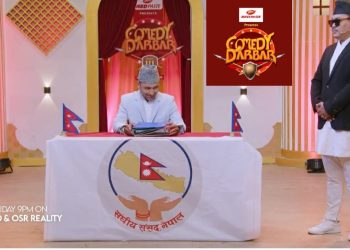 The image shows a scene from the Nepali comedy show 'Comedy Darbar'. A man dressed in a white suit and grey hat sits at a table with the logo of the Nepali parliament on it, holding some papers. Another man in a black suit, white pants, and a hat stands to his right. The setting appears to be a TV studio with a red and gold backdrop.