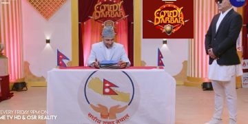 The image shows a scene from the Nepali comedy show 'Comedy Darbar'. A man dressed in a white suit and grey hat sits at a table with the logo of the Nepali parliament on it, holding some papers. Another man in a black suit, white pants, and a hat stands to his right. The setting appears to be a TV studio with a red and gold backdrop.