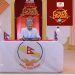 The image shows a scene from the Nepali comedy show 'Comedy Darbar'. A man dressed in a white suit and grey hat sits at a table with the logo of the Nepali parliament on it, holding some papers. Another man in a black suit, white pants, and a hat stands to his right. The setting appears to be a TV studio with a red and gold backdrop.