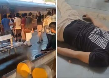 A chaotic scene at Shahdara Railway Station in Delhi, India. A crowd of people are standing near a train, while a man lies on the ground, appearing to be injured or unconscious. The man on the ground is wearing a black t-shirt with white text. In the background, a group of people are gathered, some of whom appear to be arguing or physically confronting each other. The setting is a railway station platform with a train visible in the background.