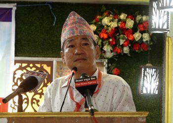 Hark Sampang Rai, Chairman of Shram Sanskriti Party, speaking at a podium with microphones from Now Army News and The News Agency, wearing a traditional Nepali hat and white shirt, in a decorated room with flowers and green wall.