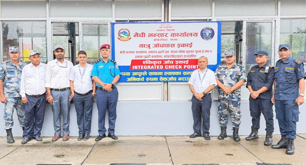 Group photo of officials from various agencies standing in front of a signboard for the Integrated Check Point at Kakarbhitta border crossing. Visible are (from left to right): a security officer in camouflage uniform, officials in plain clothes, and uniformed personnel from Nepal Police, Armed Police Force, and Nepal Customs. They are standing in front of a large sign with Nepali and English text. The scene is set on a concrete pavement outside a building with glass windows.