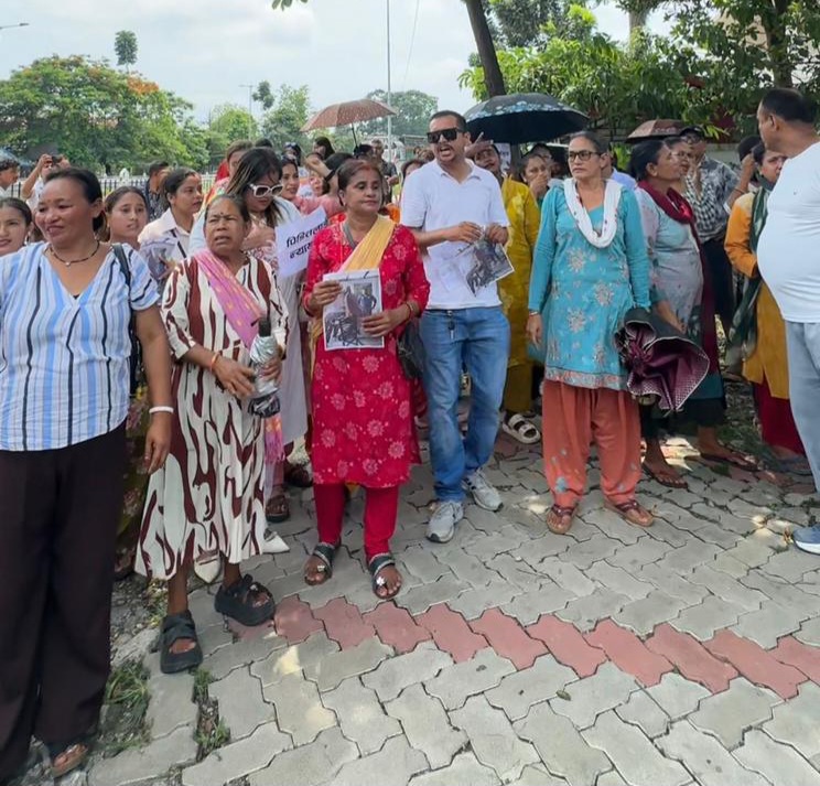 A group of people, including women in traditional clothing, are gathered on a brick-paved street, holding signs and pictures. They appear to be protesting or demonstrating, with some looking directly at the camera. The scene suggests a community event or rally, possibly related to a local issue or social cause.