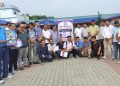 A group of approximately 25 men, mostly wearing casual clothing and some holding certificates or booklets, stand and kneel on a brick-paved surface. They appear to be participants or attendees of an event related to 'Printers Association, Jhapa' as indicated by a sign behind them. The scene is set outdoors with buildings and trees in the background under a cloudy sky.