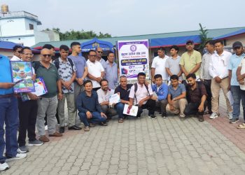 A group of approximately 25 men, mostly wearing casual clothing and some holding certificates or booklets, stand and kneel on a brick-paved surface. They appear to be participants or attendees of an event related to 'Printers Association, Jhapa' as indicated by a sign behind them. The scene is set outdoors with buildings and trees in the background under a cloudy sky.