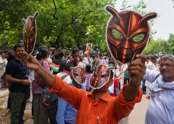 A man in an orange shirt holds up a red mask with horns covering his face, surrounded by a crowd of people, some holding signs and Indian flags, in a street with trees in the background.